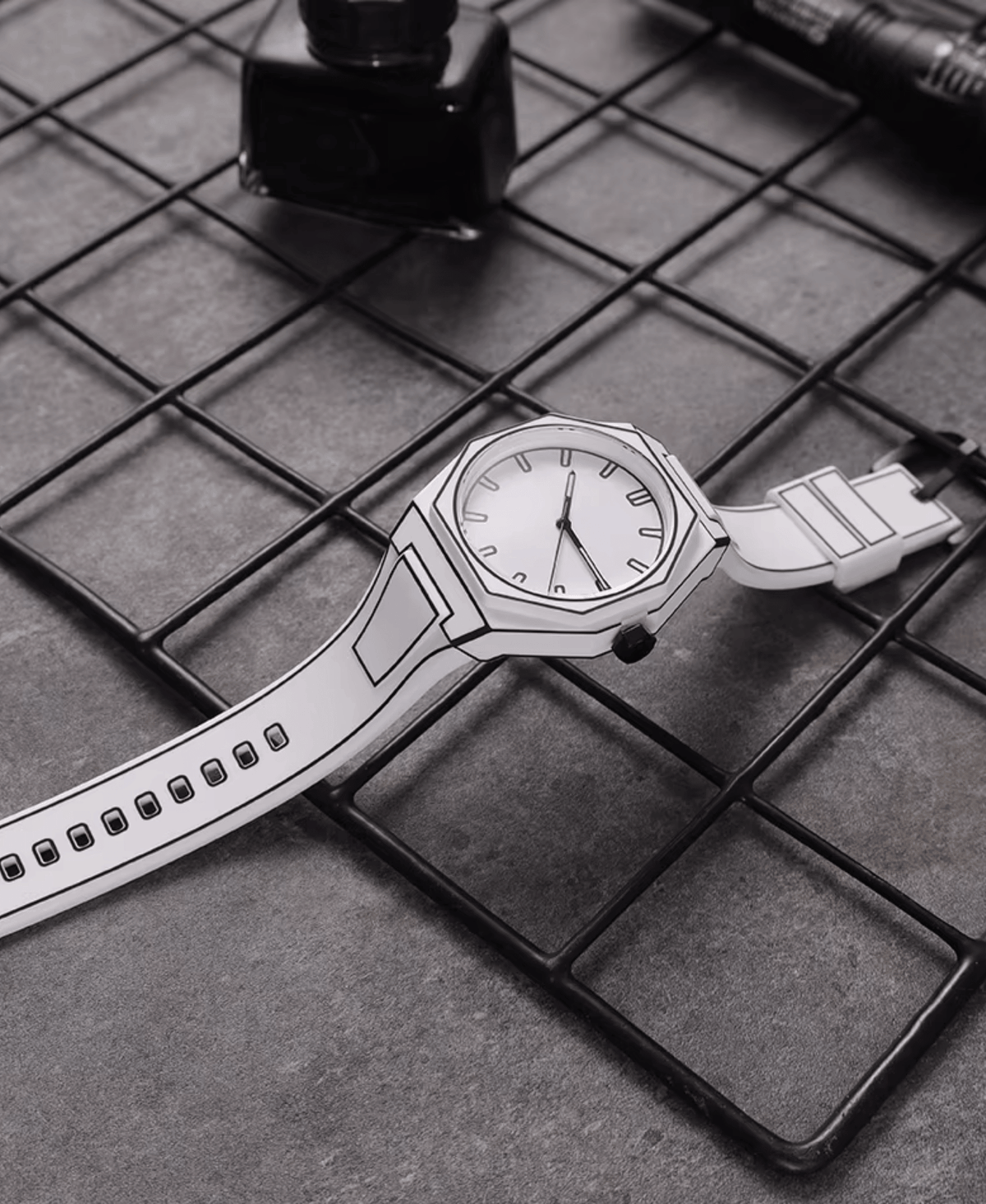 A white watch with black outline details, resembling a sketch, lying on a dark grey metal grid surface. The watch has a white face, black hour markers, and a white strap. An ink bottle and a pen are visible in the blurred background.