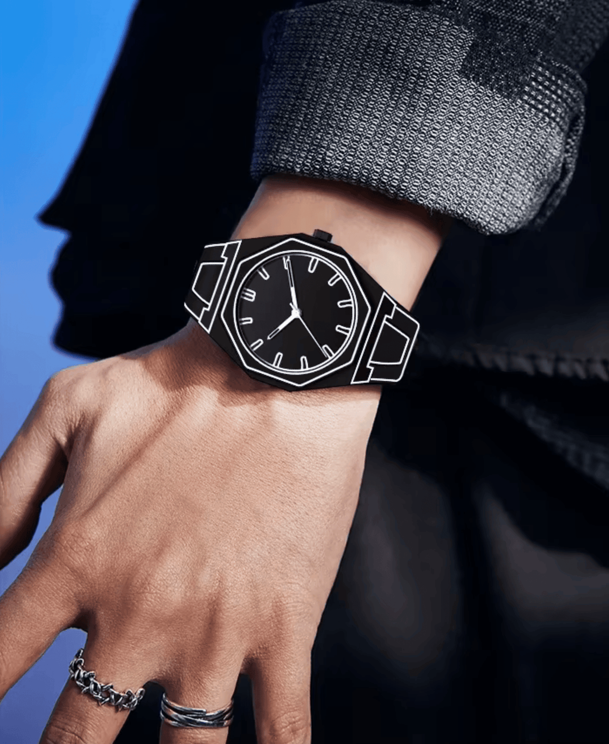 Close-up of a man's hand wearing a black watch with white outline details. The watch has a minimalist design with a black face and white hour markers. The man is wearing a dark grey jacket and silver rings on his fingers. The background is a dark blue gradient.